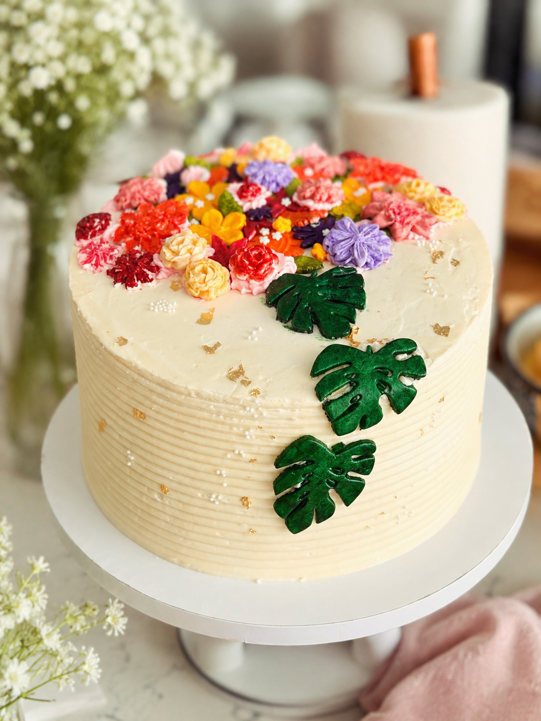 Ivory birthday cake topped with colorful buttercream flowers and green monstera leaves